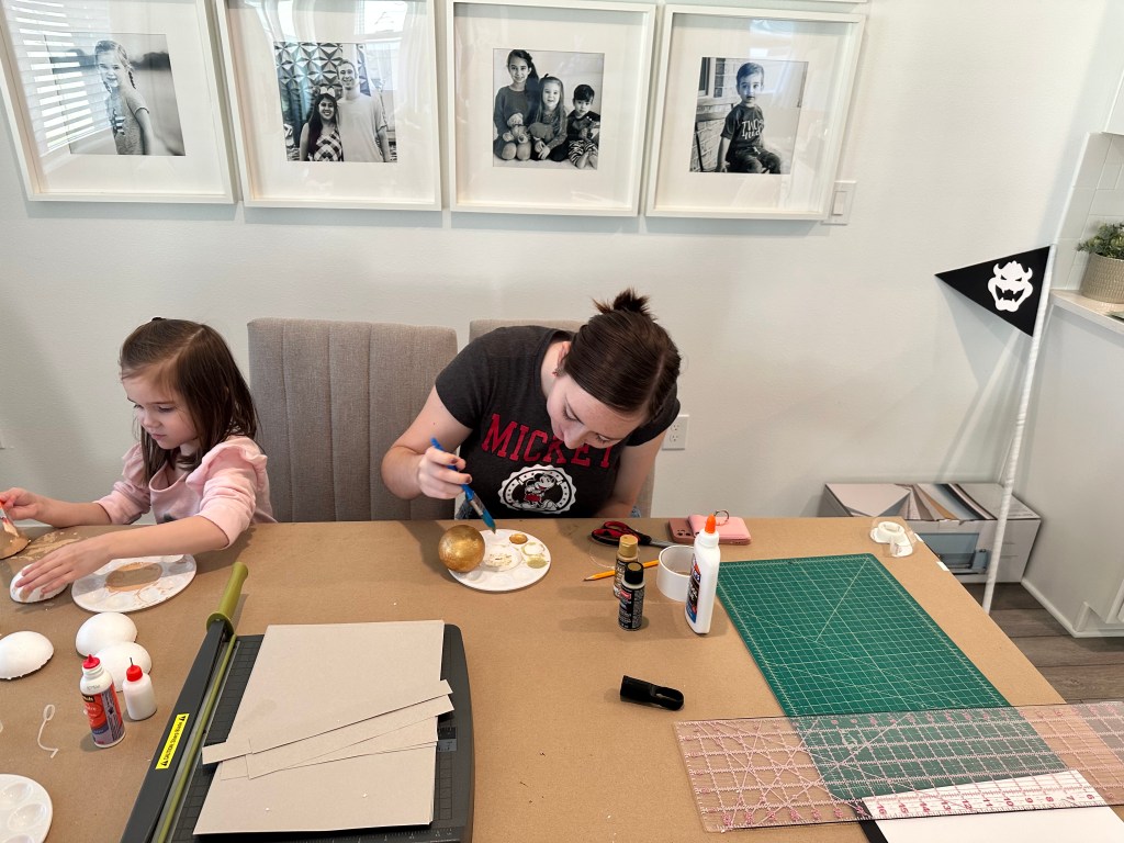 A young girl painting a gold foam ball for a Mario birthday party Bowser’s flag project, sitting at a craft table with supplies and tools.