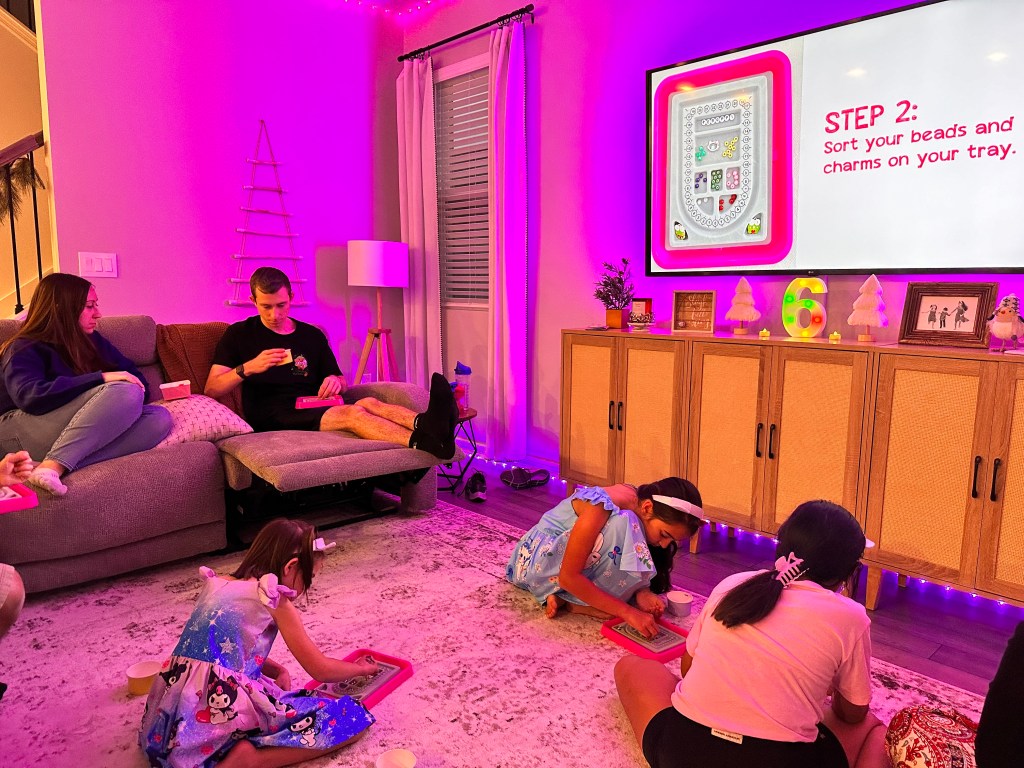 Guests seated in a living room watching a screen showing bracelet-making steps while sorting beads on pink trays.