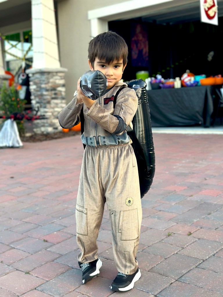 Young boy in a Ghostbusters costume holding an inflatable proton blaster in front of a decorated garage on Halloween.
