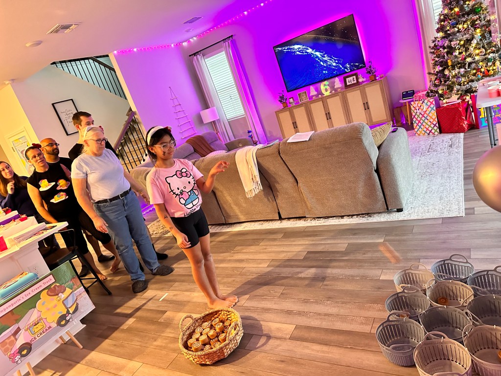 Guests lined up behind a girl tossing a felt cinnamon roll toward a set of baskets in a decorated living room.