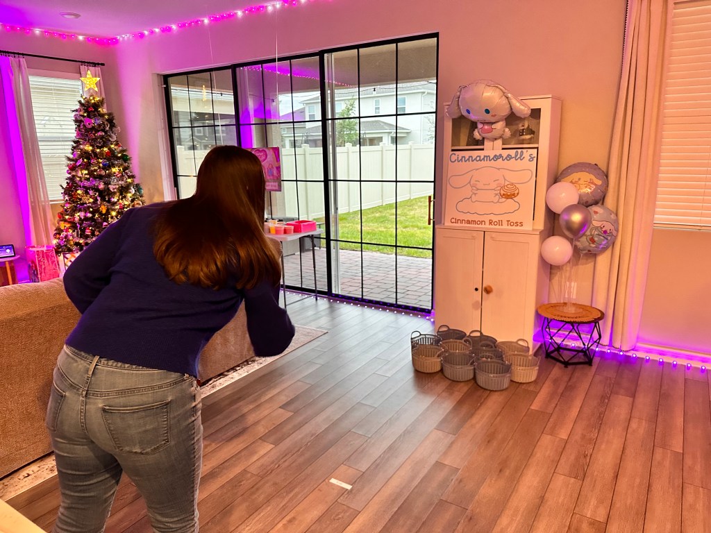 A woman tossing a felt cinnamon roll toward baskets under a Cinnamoroll sign in a festively lit living room.