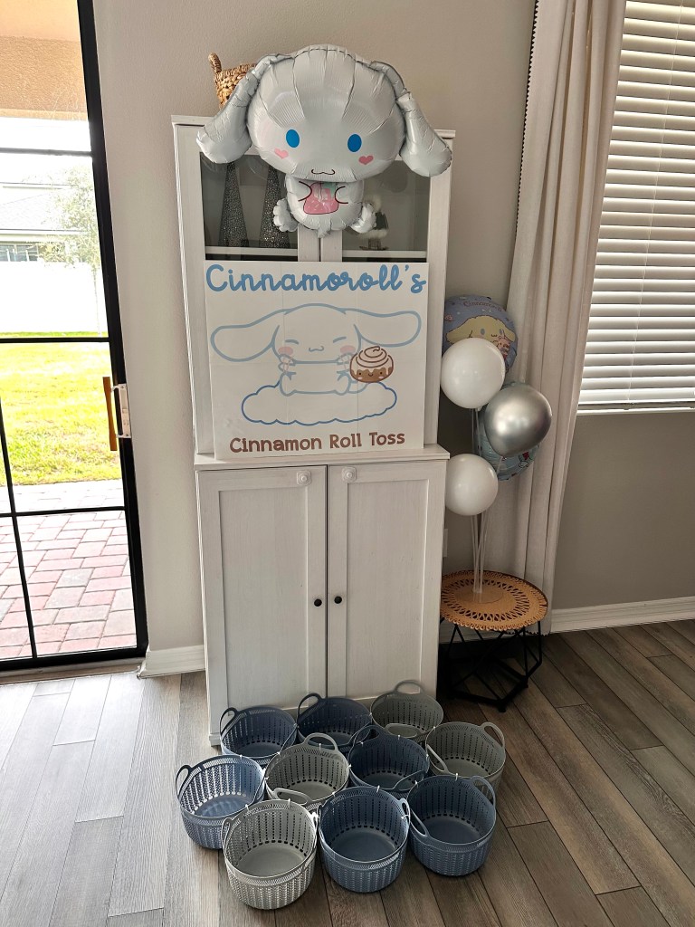 A party game station featuring a Cinnamoroll sign, a Cinnamoroll balloon, pastel-colored baskets arranged on the floor, and a stack of cinnamon roll toss items.