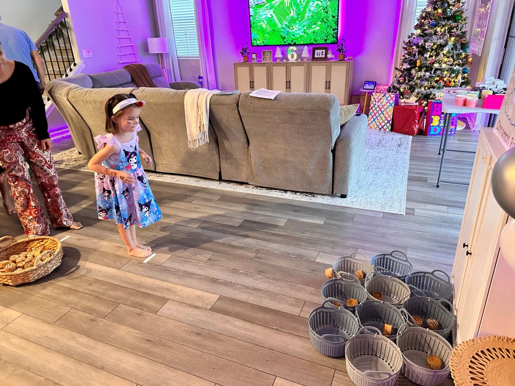 Young girl in a Kuromi dress tossing a felt cinnamon roll toward baskets lined up on the floor at a Sanrio-themed birthday party.