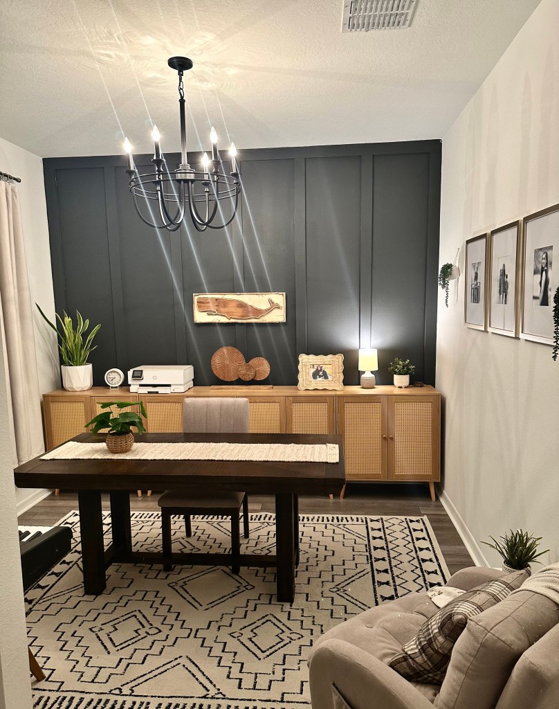 Den and craft room setup before party decorations, featuring a table, rug, wall art, and shelving with plants and decor items.