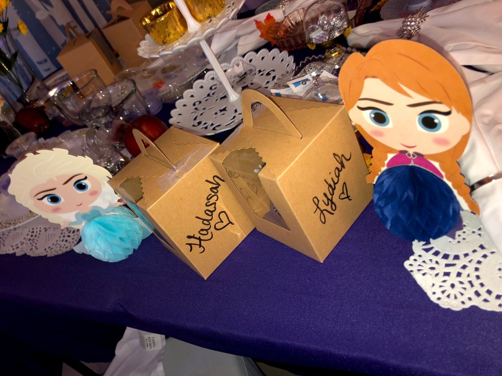 Two brown cupcake boxes labeled with names sit next to Elsa and Anna paper decorations on the table, ready to carry home the girls’ creations.