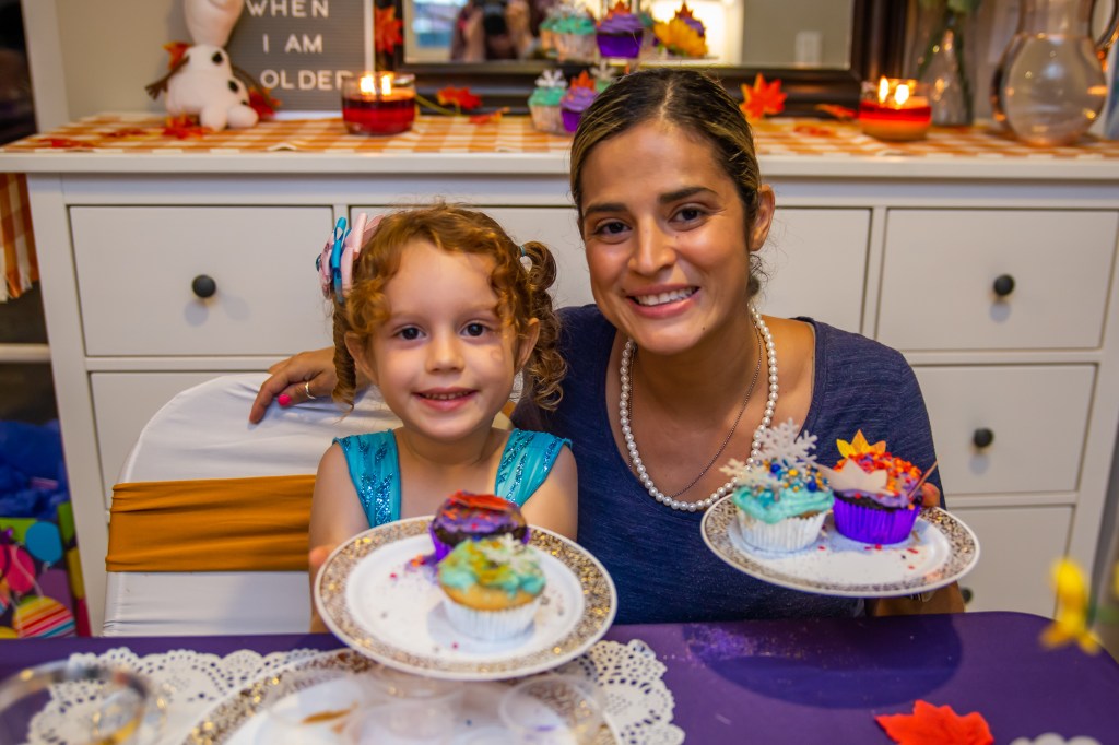 A girl and her mom pose proudly with their completed cupcakes, featuring snowflake and leaf toppers, in front of a fall-themed dessert table.