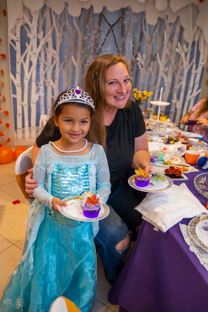 A young girl in an Elsa dress smiles beside her mom, both holding plates with beautifully decorated cupcakes inspired by Frozen 2.