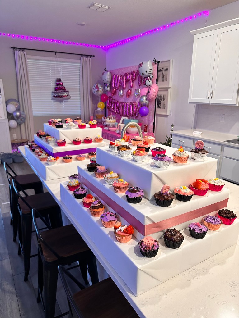 Large tiered cupcake stands on a kitchen island filled with pink, purple, and chocolate cupcakes from the Cupcake Wars activity.