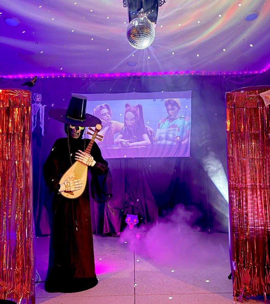 A dramatic entrance to the Demon World featuring fog effects and metallic curtains.  Jinu, dressed in a skeleton robe and gat, stands in front playing a bipa, while a projection of the K-Pop Demon Hunters plays in the background. Derpy Tiger peeks out beneath the screen.