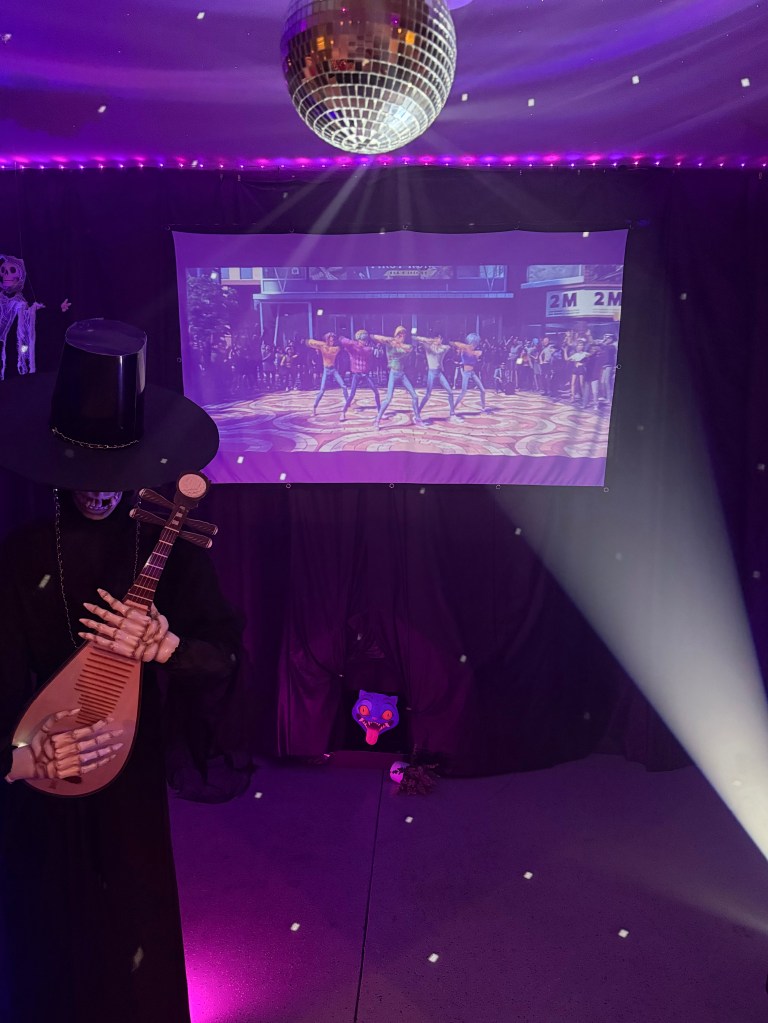 Disco ball reflecting light beams across the Halloween garage as a spotlight shines upward. Below, the K-Pop Demon Hunters dance video plays on the projected screen surrounded by black curtains, purple LED lights, and spooky props including a skeleton musician and hidden Derpy Tiger