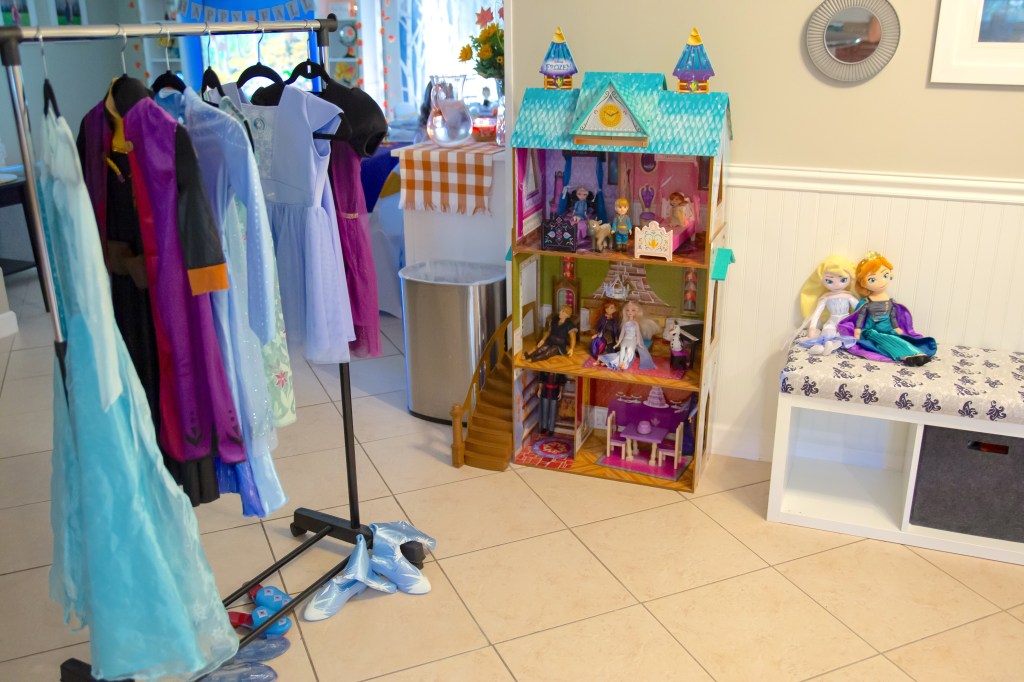 Dress-up corner with Frozen costumes on hangers, princess shoes on the floor, and a large Frozen dollhouse nearby.
