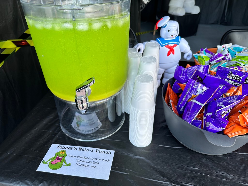Close-up of a drink dispenser filled with neon green punch next to stacked cups, a Stay Puft Marshmallow Man toy, and bowls of Halloween snacks.