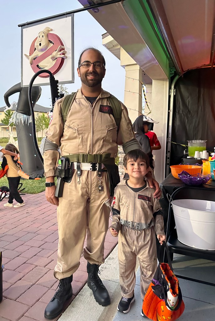 Adult and child dressed in matching Ghostbusters jumpsuits smiling outside a decorated garage, holding a Halloween trick-or-treat bag.