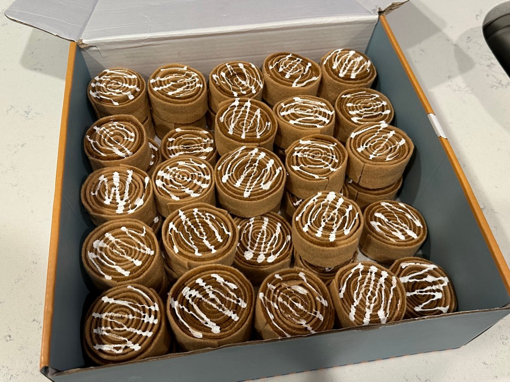 A box filled with rolled brown felt circles decorated with white icing squiggles, resembling cinnamon rolls for a party game.