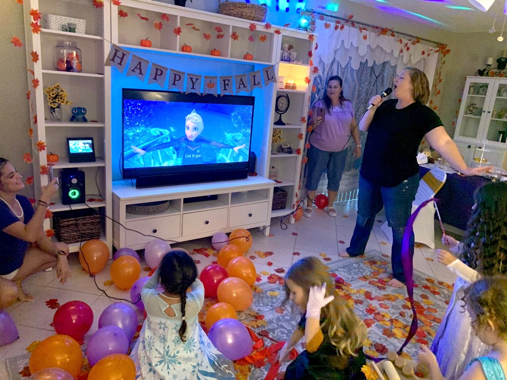 A mom enthusiastically sings into a microphone while several kids join in with ribbon dancing and leaf tossing. The room glows with colorful lights, and the TV displays the Frozen karaoke scene for “Let It Go.”