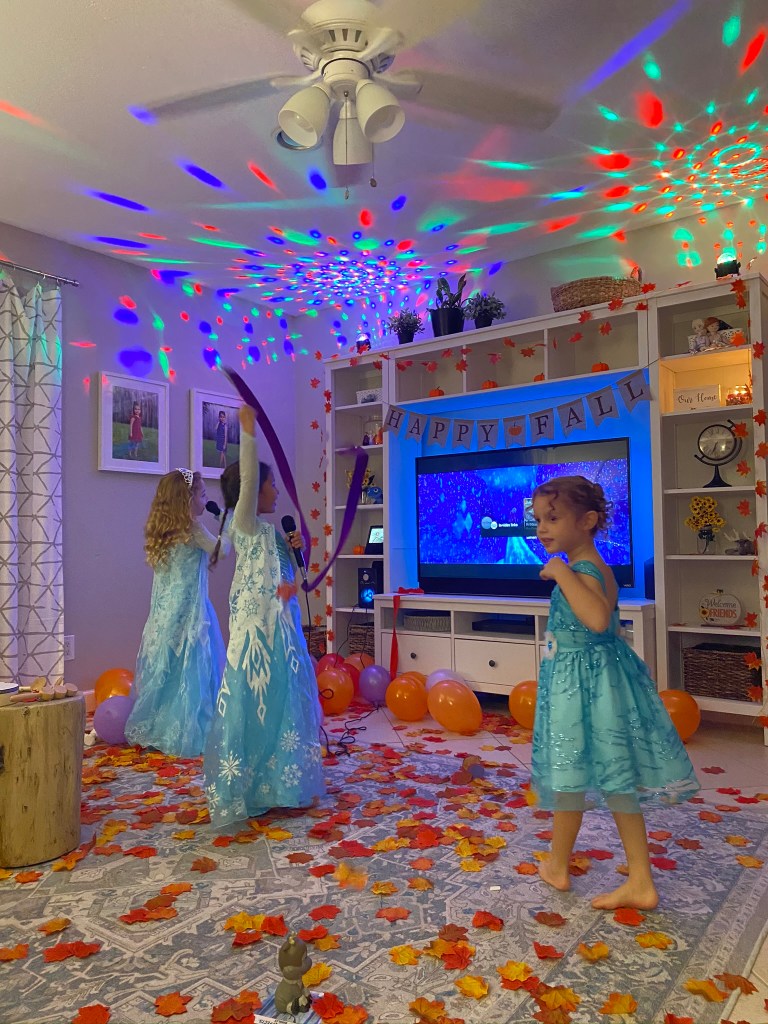 Three girls in Frozen costumes sing and dance in a living room decorated with autumn leaves, balloons, and disco lights, with a Frozen karaoke video playing on the TV.