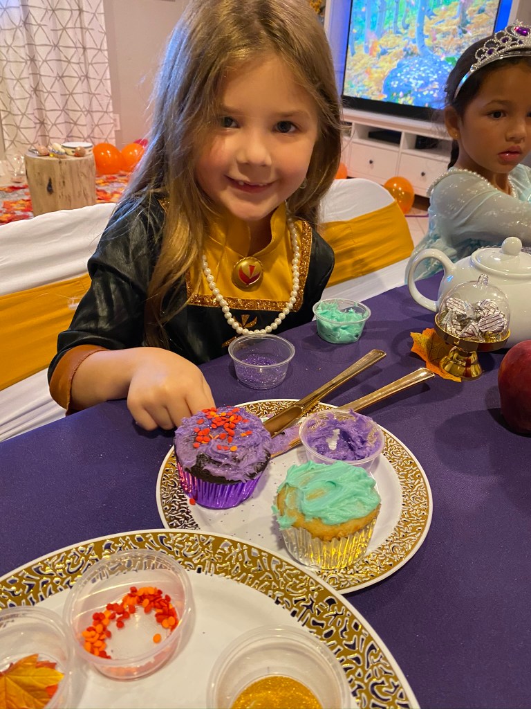 A smiling girl in an Anna costume proudly shows off her cupcakes topped with purple frosting and orange heart-shaped sprinkles.