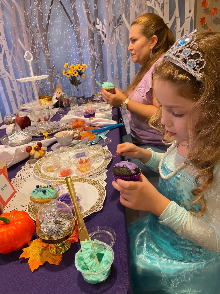 A young girl in an Elsa dress and tiara carefully spreads frosting onto a chocolate cupcake while decorating with colorful sprinkles and themed toppings.