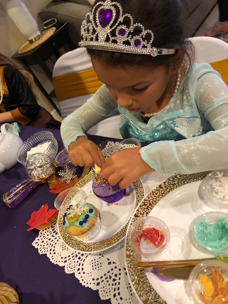 A girl in an Elsa costume with a purple jeweled tiara decorates her cupcakes with purple frosting, rainbow sprinkles, and fall leaves while a finished cupcake with blue pearls and a snowflake topper sits nearby.