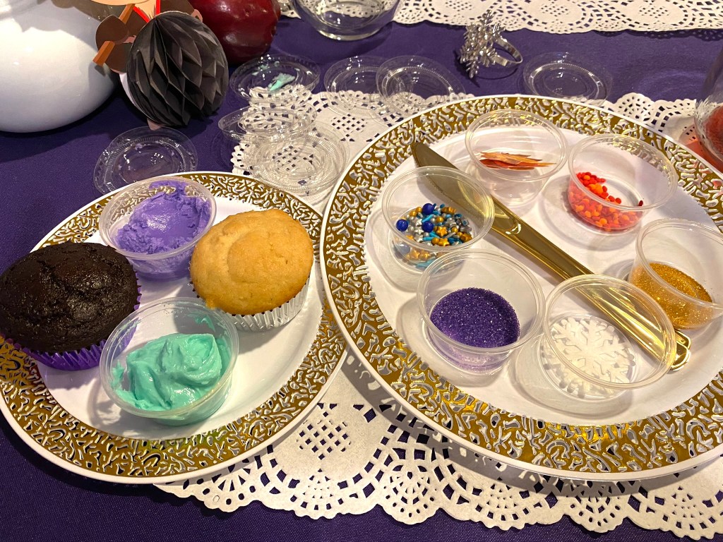 Two unfrosted cupcakes on a gold-trimmed plate with purple and aqua frosting, surrounded by individual cups of sprinkles, sugar crystals, snowflakes, and edible pearls for decorating.