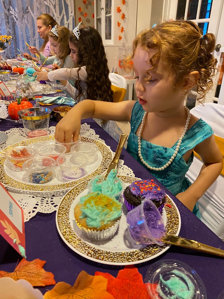A young girl in a blue Frozen dress reaches for sprinkles as she decorates two cupcakes with swirled aqua and purple frosting, gold shimmer, and orange heart confetti.