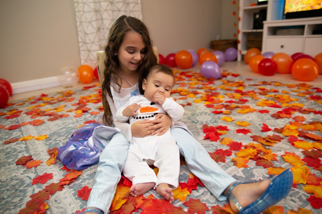 Elsa laughs while holding baby Noah on her lap, dressed as Olaf, as he sucks on his hand amid a whimsical backdrop of fall leaves and colorful party balloons.