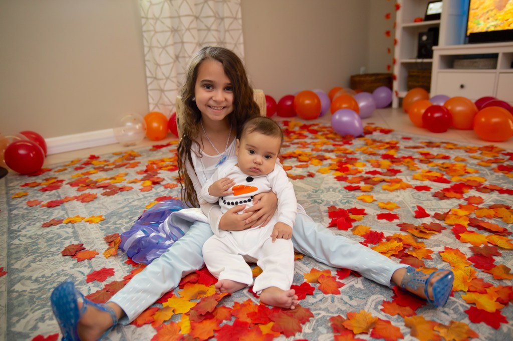 Elsa smiles at the camera while holding her baby brother Noah, who is dressed as Olaf the snowman, surrounded by scattered autumn leaves and balloons on a cozy living room rug.