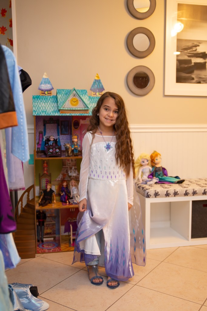 Birthday girl dressed as Elsa stands smiling beside her Frozen dollhouse before the party begins.