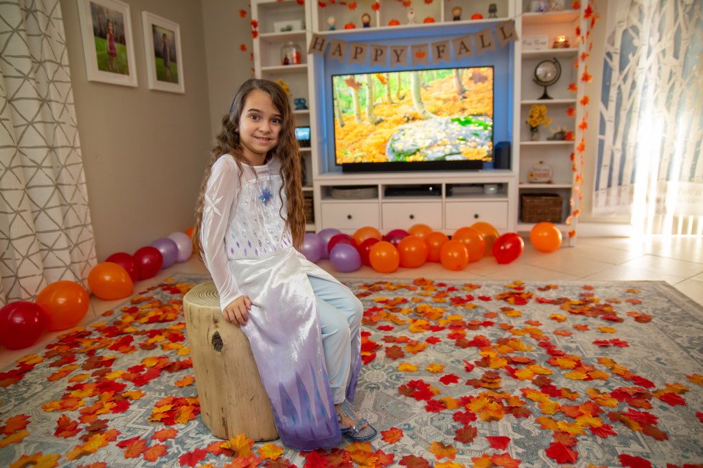Birthday girl sits on a log-style stool surrounded by fall leaves and balloons, with a Frozen forest scene playing on TV.