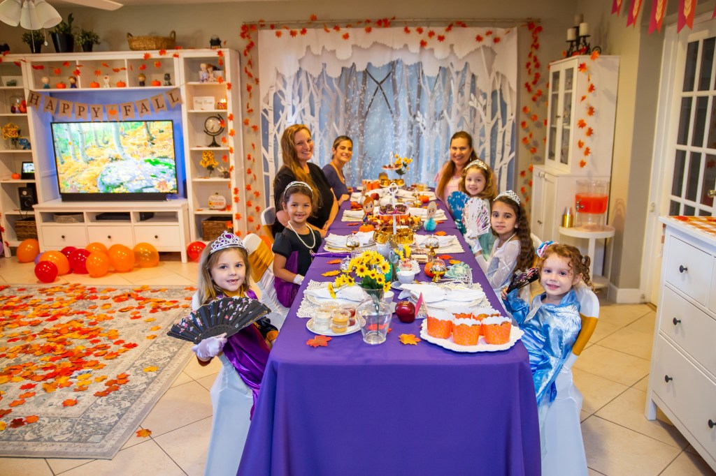 A wide shot of the full Frozen 2 tea party setup, showing young guests dressed as princesses and several smiling moms seated at a long, richly decorated table with fall and Frozen-themed details.