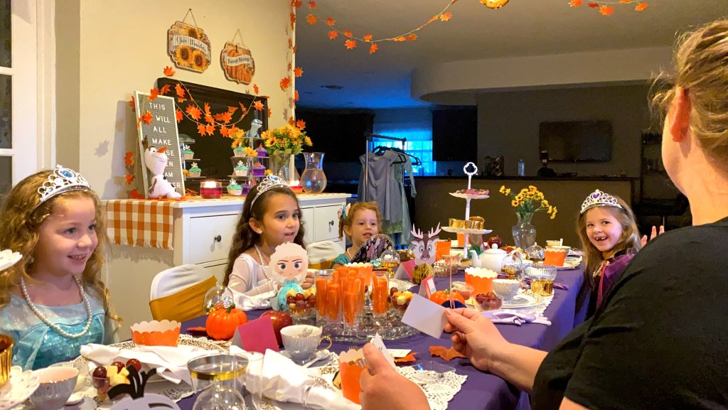 A group of young girls dressed as princesses sit at a Frozen-themed tea party table, smiling as one of the moms reads lyric clues during a “Guess the Lyrics” game. The table is decorated with Frozen characters, autumn leaves, snacks, and teacups.