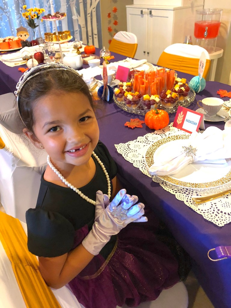 A smiling young girl dressed in a tiara, pearls, and princess gown sits at the Frozen-themed tea party table, with festive snacks and place settings arranged in front of her.