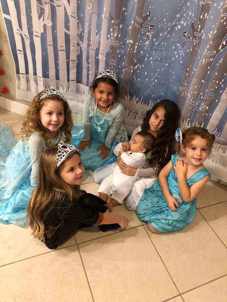 A group of young girls in Frozen-themed princess dresses gather around baby Noah, who is dressed as a snowman. They are seated in front of a birch forest Frozen 2 backdrop, smiling and looking at him affectionately.
