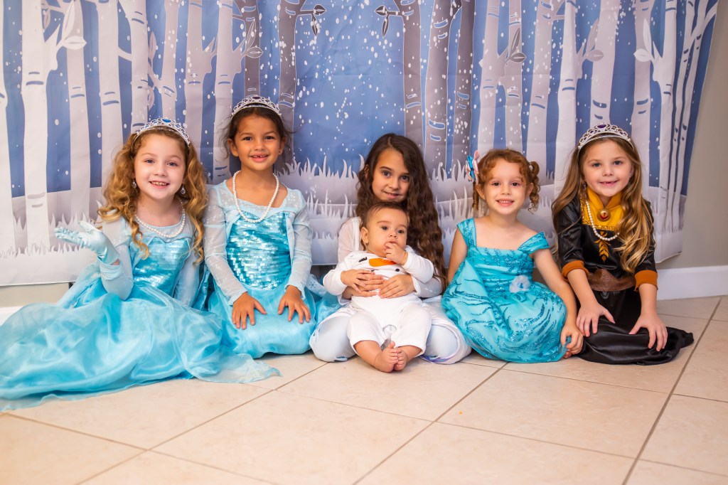 Six children dressed as Frozen characters pose together in front of a snowy birch tree backdrop. In the center, Elsa holds her baby brother Noah, who is dressed as a snowman. Everyone is smiling and looking at the camera.