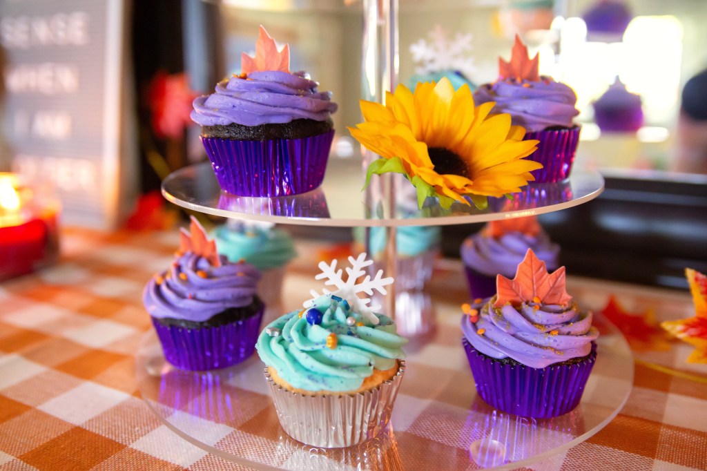 Close-up view of Frozen 2–themed cupcakes on a tiered stand. The purple cupcakes are decorated with edible fall leaves to represent Anna, and the light blue cupcakes are topped with white snowflakes and shimmering sprinkles to represent Elsa.