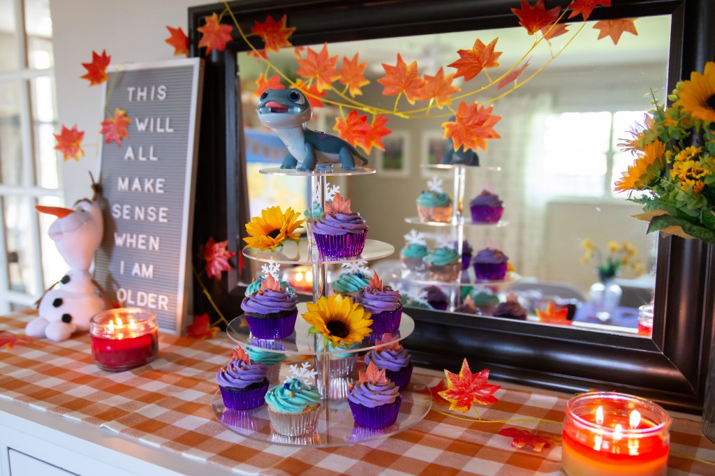 Cupcake display on a checkered tablecloth with a tiered stand holding purple and teal cupcakes, topped with snowflakes and leaves. Olaf plush stands next to a letter board sign with fall leaves and candles.