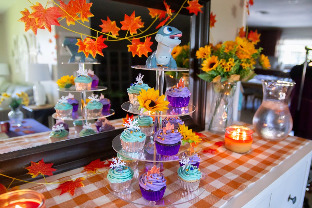 Tiered cupcake stand decorated with teal and purple cupcakes, fall leaves, and snowflakes. At the very top sits a Bruni the Fire Spirit figurine from Frozen 2, surrounded by a sunflower and backed by autumn leaves.