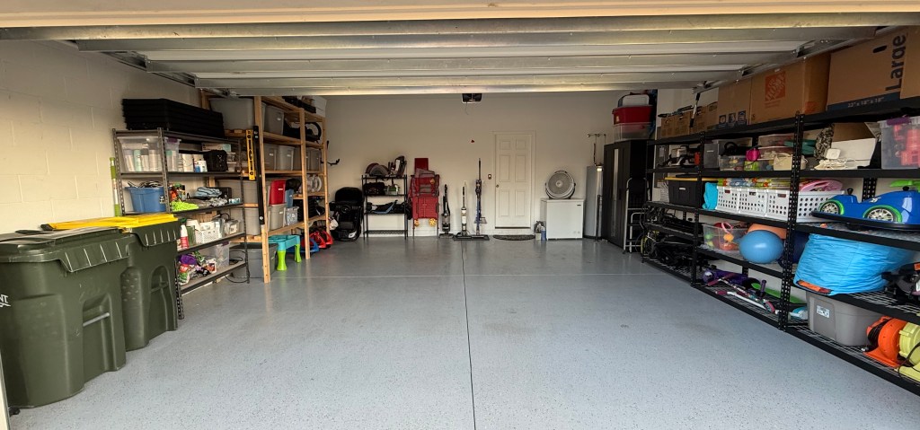 Two-car garage with shelves full of storage bins, cleaning supplies, kids’ toys, and vacuum equipment—captured before transforming the space into a Halloween setup.