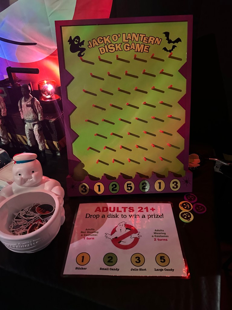 Close-up of a neon green and purple Jack-O’-Lantern themed plinko game board for adults, with Stay Puft bowl full of disks and prize chart for winning jello shots or candy.