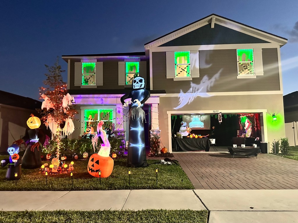 A two-story home decorated for Halloween with green lighting, inflatables, glowing ghosts projected on the house, and a garage transformed into a Ghostbusters-themed candy station.