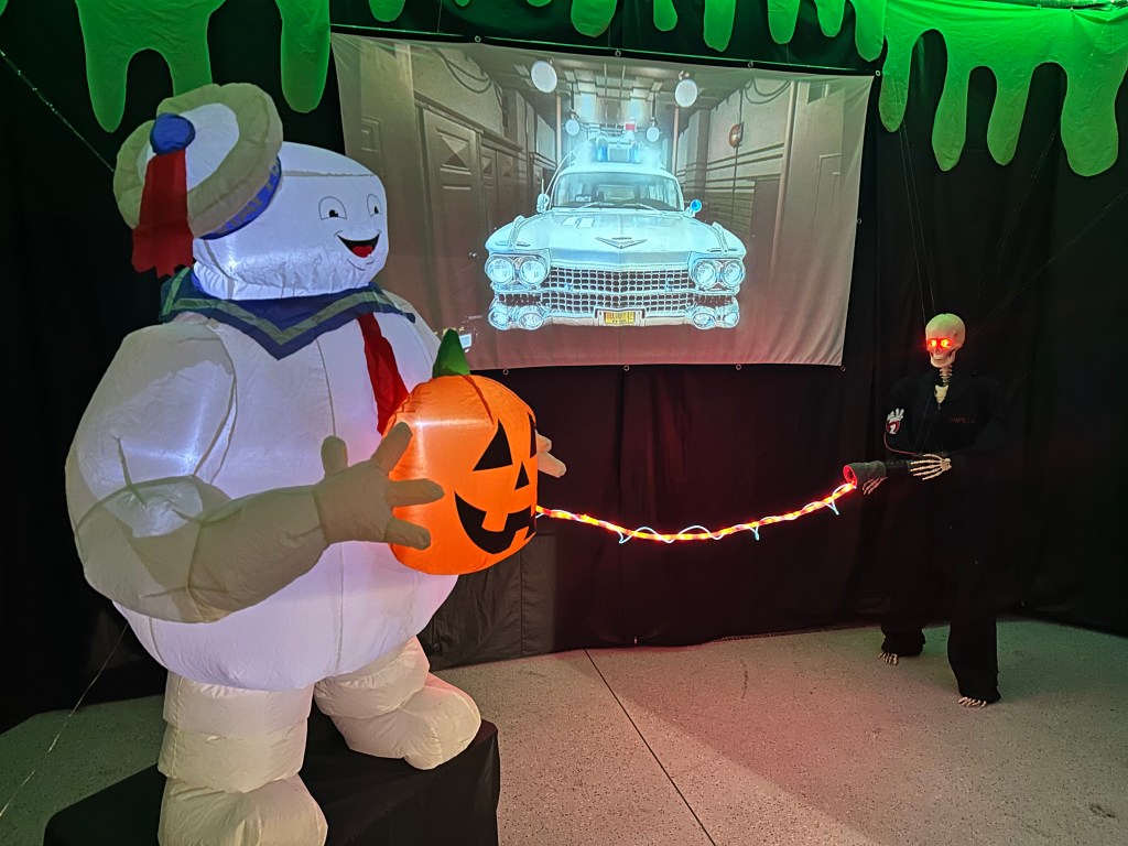 Inflatable Stay Puft Marshmallow Man holding a glowing jack-o’-lantern with a projected image of the Ecto-1 and a glowing-eyed skeleton Ghostbuster shooting a red LED proton beam