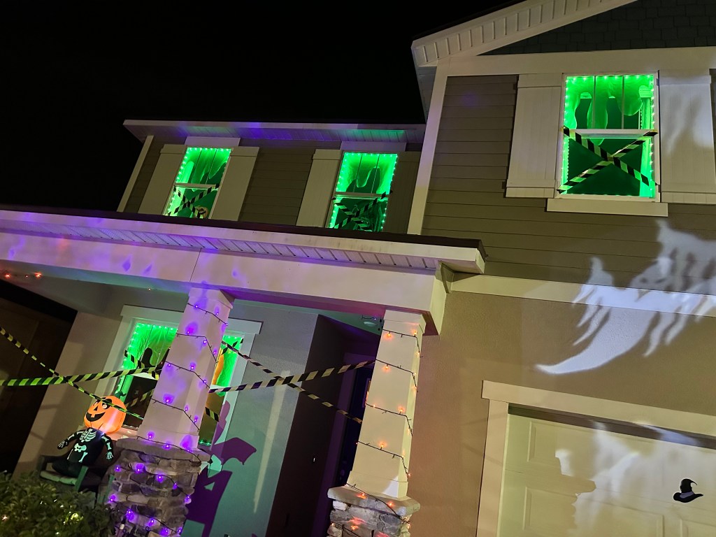 Close-up of Halloween house windows lit with green lights, showing ghost silhouettes and X-pattern caution tape.