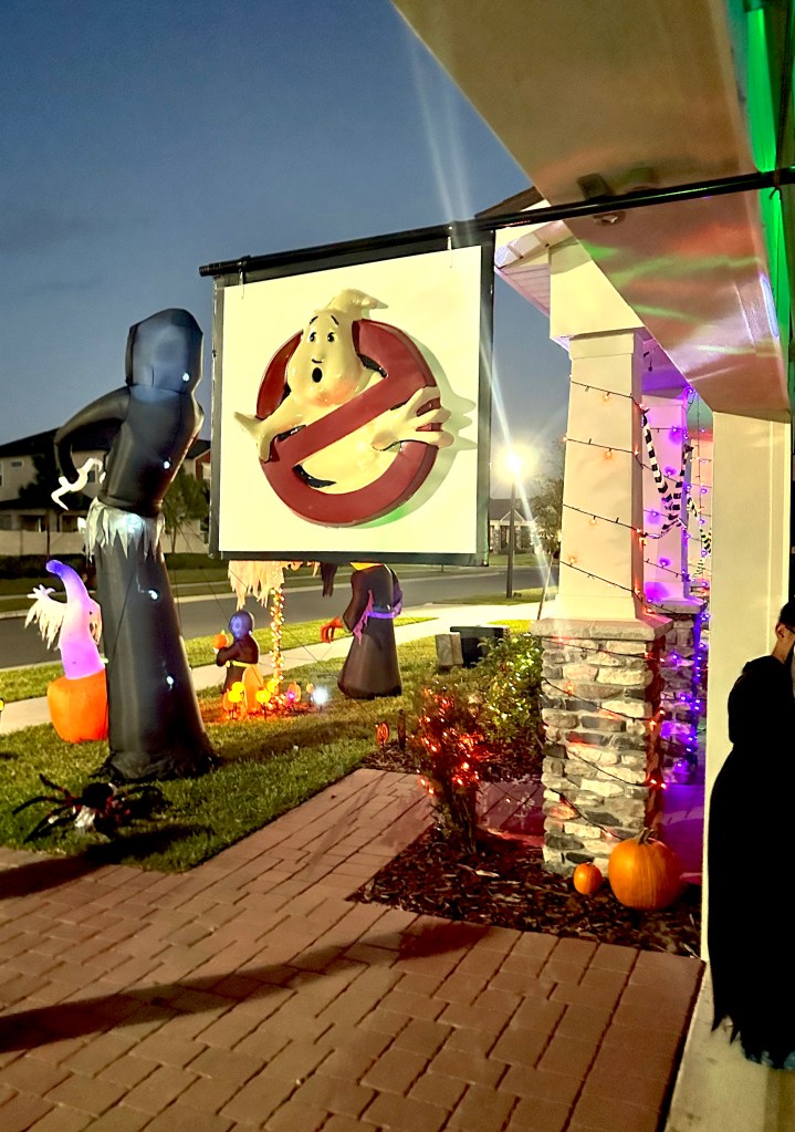 Handcrafted Ghostbusters sign with 3D logo hanging from a suburban garage, surrounded by Halloween inflatables and purple-orange lights.