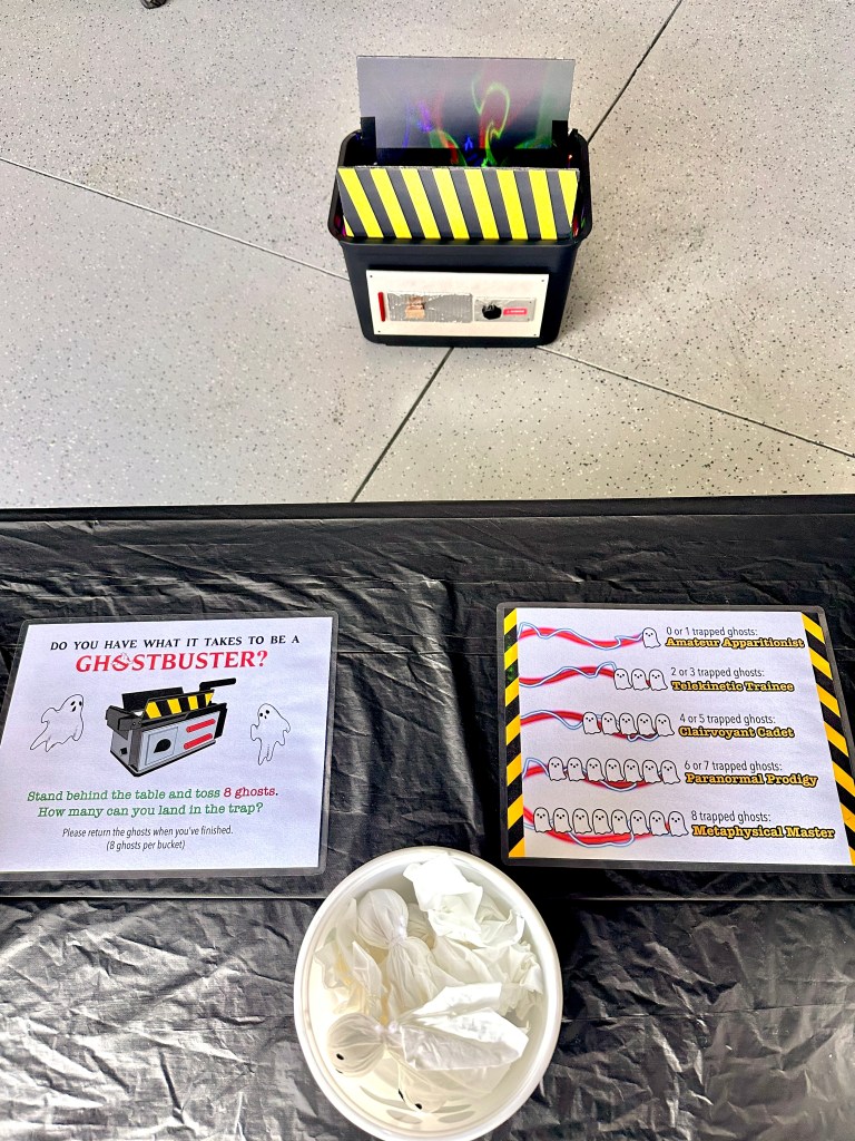 Ghostbusters Halloween game station with ghost trap bucket, toss instructions, ghost rank chart, and bowl of DIY tissue ghosts.