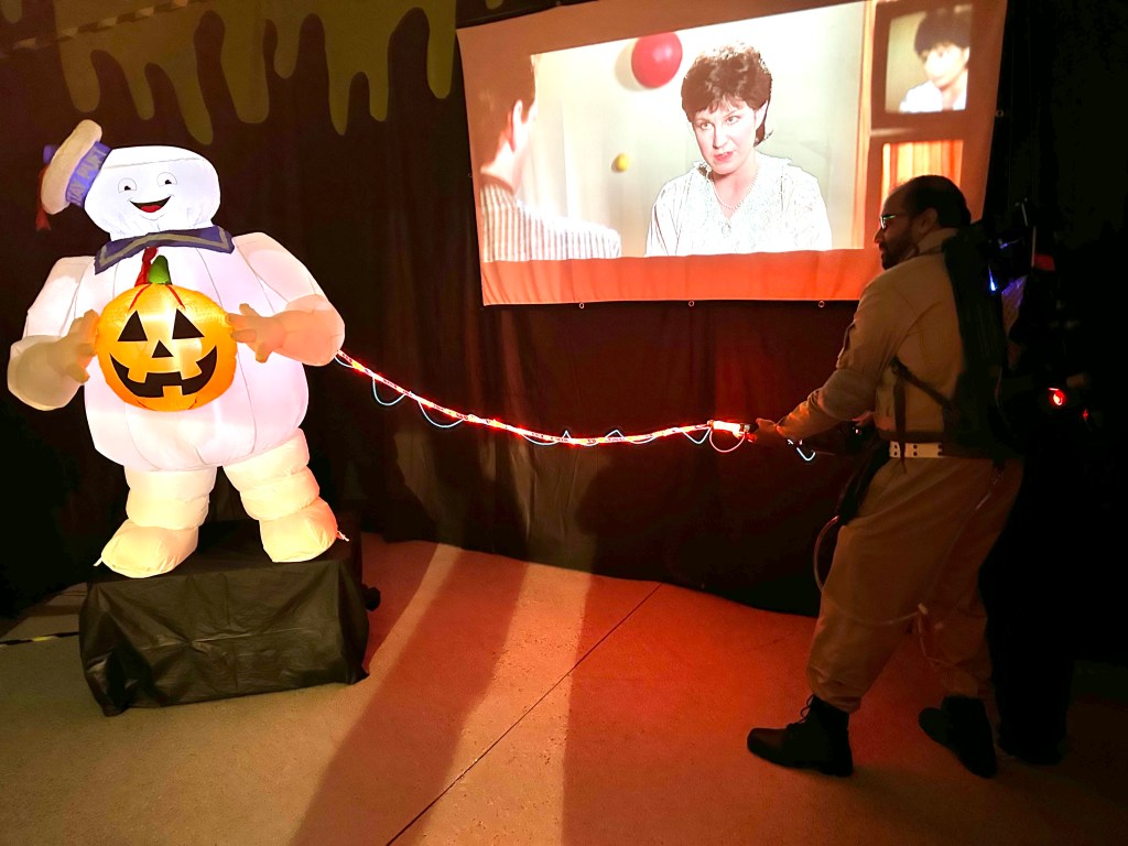 Man in Ghostbusters costume aiming a red glowing proton blaster at an inflatable Stay Puft figure while a scene from the movie plays on a projector.