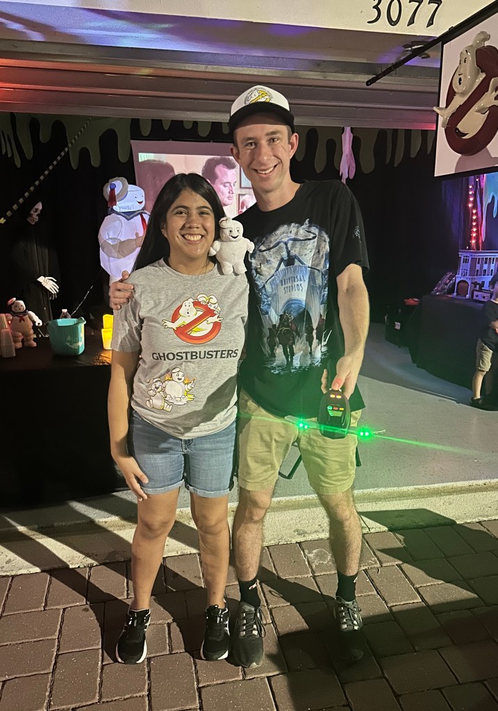 Young man and woman smiling in front of a Ghostbusters-themed Halloween garage, wearing Ghostbusters T-shirts and holding a Stay Puft plush and PKE meter with glowing green lights.