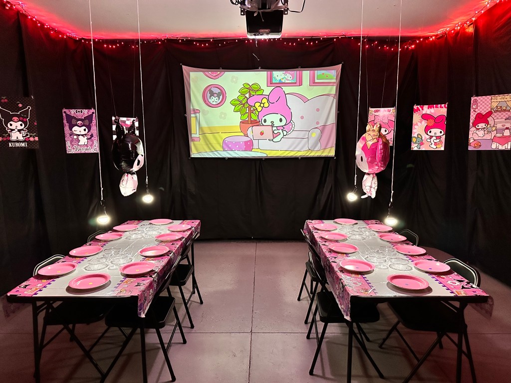 Wide view of a Hello Kitty themed garage with black drape walls, pink lights, a My Melody screen, and two decorated tables set for a cupcake decorating contest.