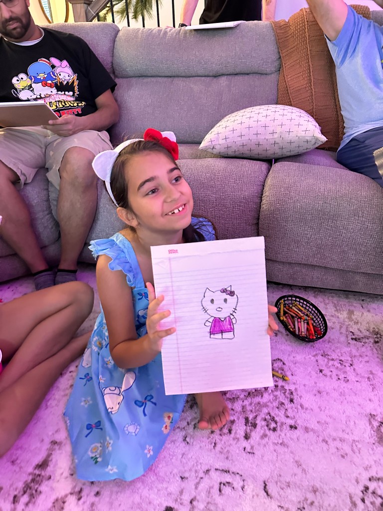 A smiling girl in a blue Hello Kitty dress holds up her completed drawing of Hello Kitty colored in purple and pink.