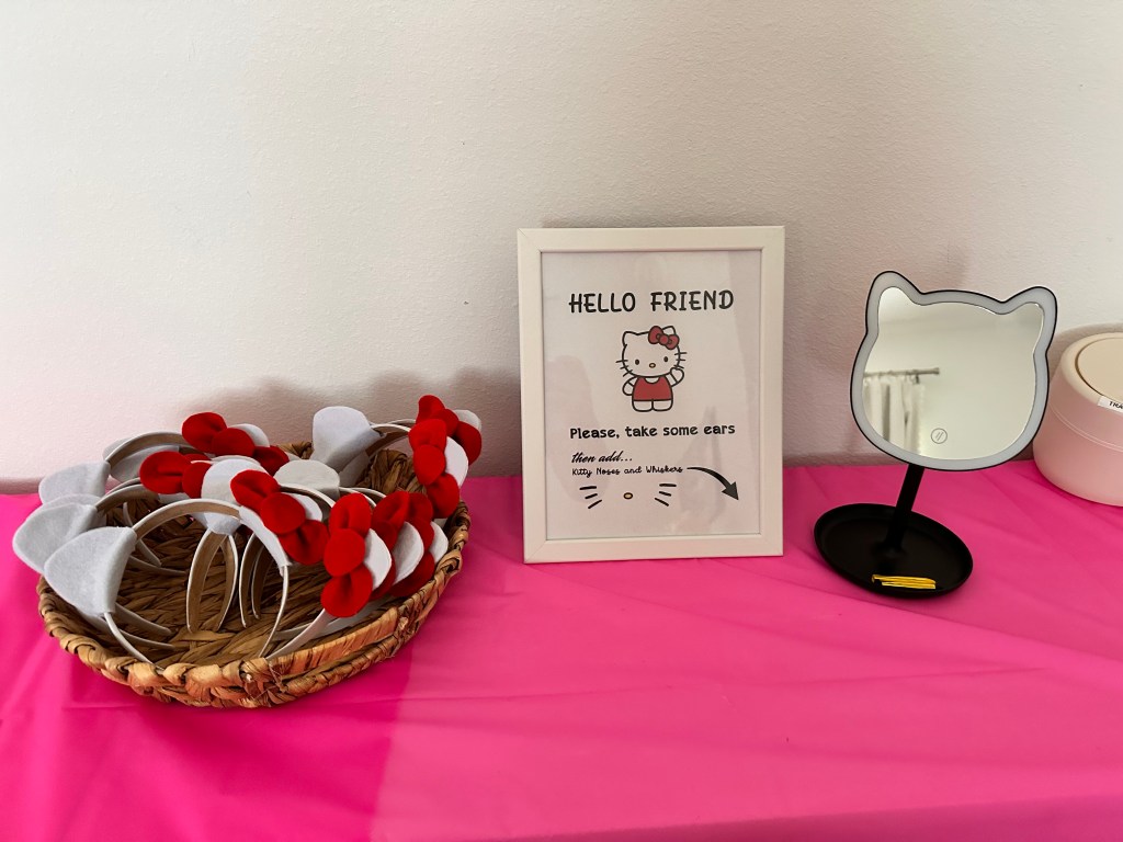 A party station with a framed Hello Kitty sign, a wicker basket of red-bow headbands, and a Hello Kitty-shaped mirror on a pink table.