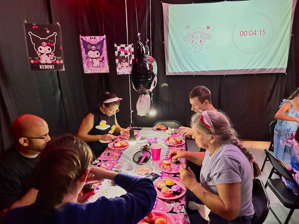Adults gathered around a table decorated with Kuromi decor, focused on decorating cupcakes while a countdown timer projects on the wall during a Hello Kitty cupcake wars game.